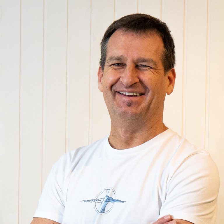 Gründer Mann mit kurzem Haar und lächelndem Gesicht, in weißem T-Shirt vor hellem Hintergrund. Bild vom Gründer der NKP.