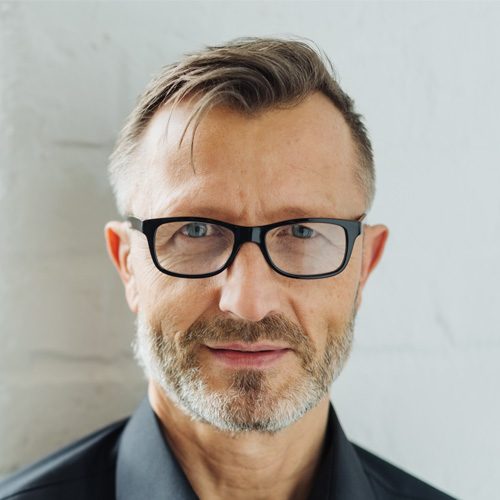 Testimonial 1 Mann mit Brille, kurzem, grauem Haar und einem ernsten Gesichtsausdruck in einem hellen Raum.