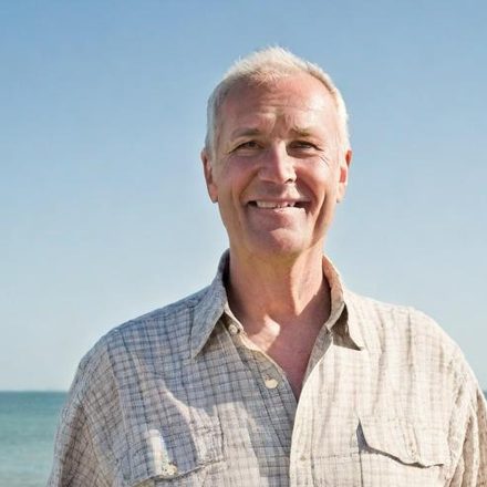 Testimonial 3 Lächelnder älterer Mann am Strand mit blauem Himmel im Hintergrund.